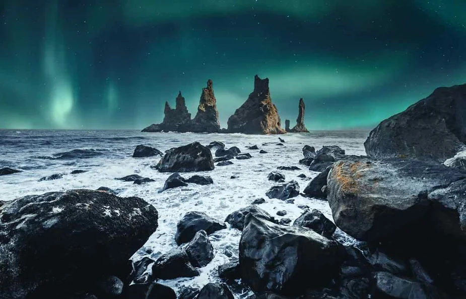 Mesmerizing view of Reynisdrangar sea stacks under the Northern Lights at Reynisfjara Beach in Iceland, with dark volcanic rocks and the Atlantic Ocean waves.