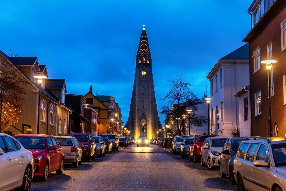 Street view in Reykjavik, Iceland, with cars parked along the sides and the illuminated Hallgrímskirkja church tower in the background during the evening.