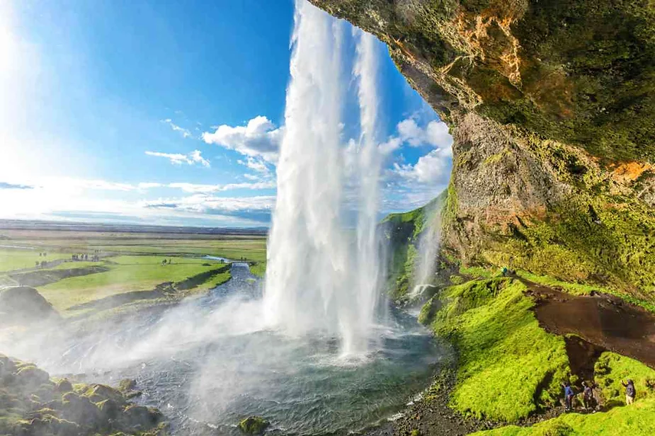 View from behind a majestic waterfall cascading down from a rocky cliff, with sunlight illuminating the water spray and lush green landscape below.