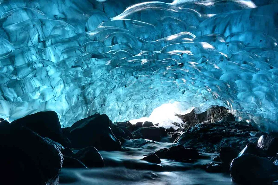 Ice cave interior with blue hues and ice formations above a rocky floor.