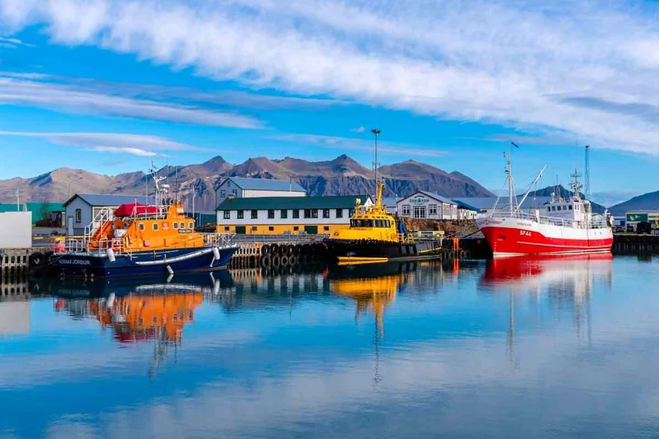 Colorful fishing boats docked at a calm harbor with mountains in the background and clear blue skies.