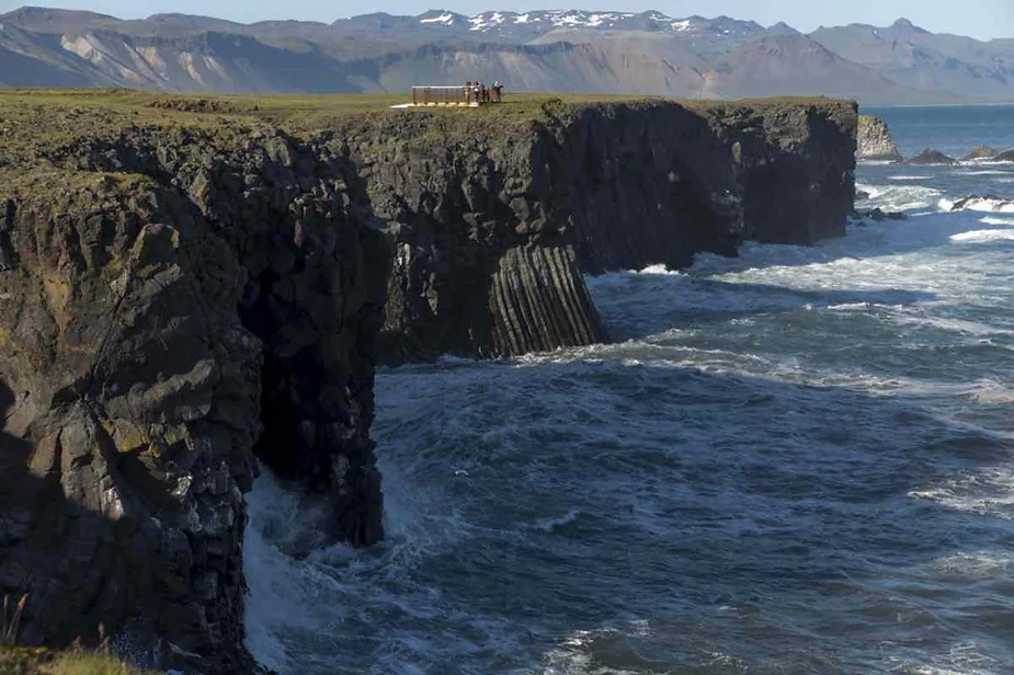 Rocky cliffs along the coastline with ocean waves crashing against the base, mountains visible in the background.