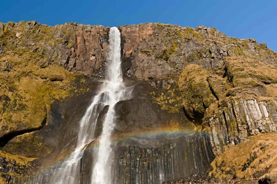 Scenic waterfall cascading down a rugged cliff with a rainbow visible at the base, under a clear blue sky.