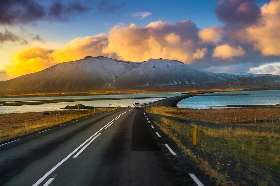 A car driving on a long, winding road with snow-capped mountains in the background, surrounded by water and illuminated by a vibrant sunset with colorful clouds.