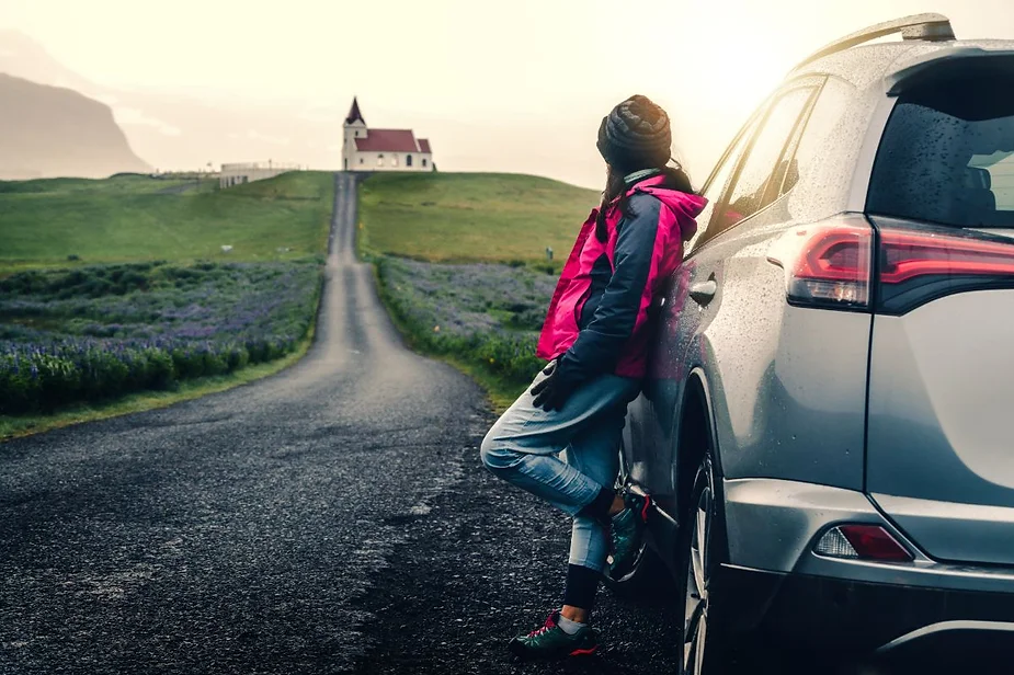 Person leaning against a silver car, gazing at a distant church at the end of a long road through green fields.