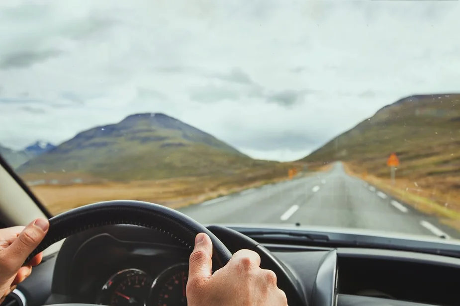 View from inside a car with a person’s hands on the steering wheel, driving on an open road surrounded by mountains.