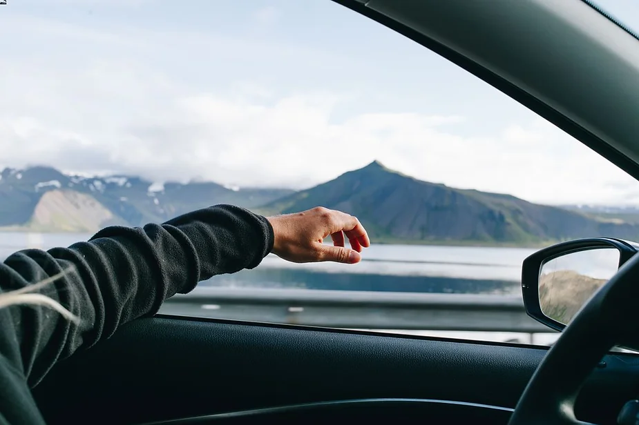 Person's arm resting out of a car window with a scenic view of mountains and a lake in the background.