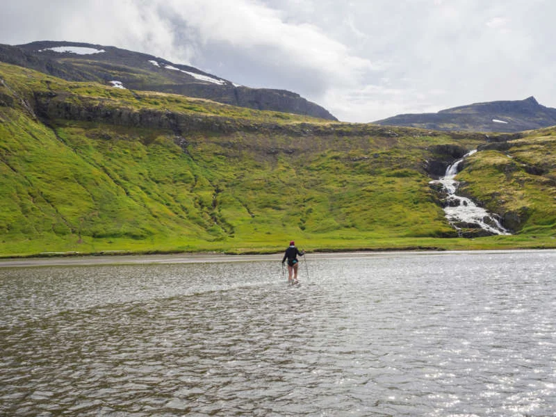 Hiker crossing a shallow river with a waterfall and green hills in the background under a cloudy sky.