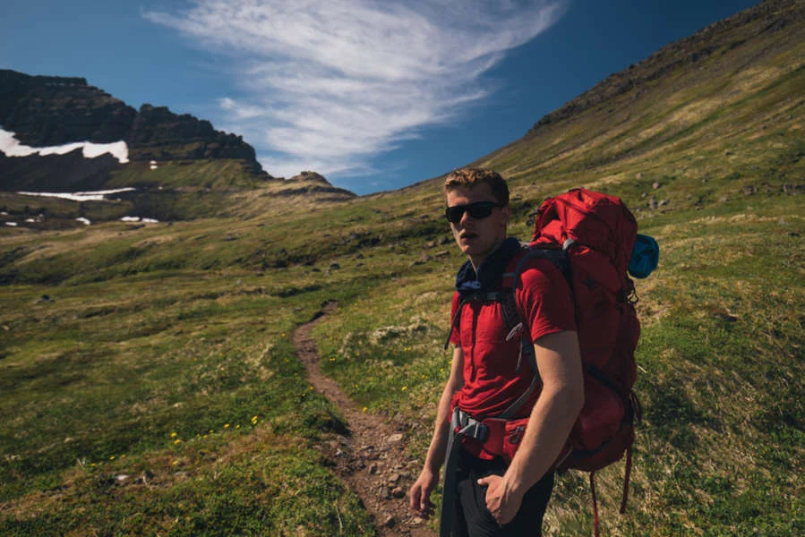 Male hiker wearing sunglasses and a red backpack standing on a grassy mountain trail under a blue sky.