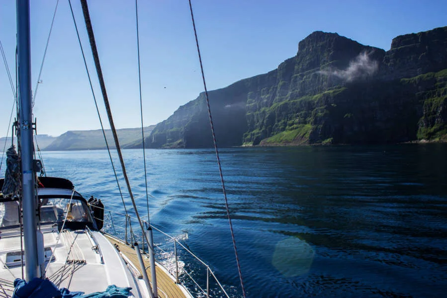 Sailing boat on calm waters near towering green cliffs under a clear blue sky.