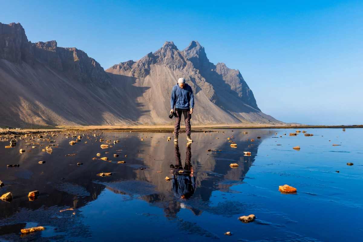 A man wearing a camera and blue jacket is reflected on the black sand beach in front of the Vestrahorn mountain.