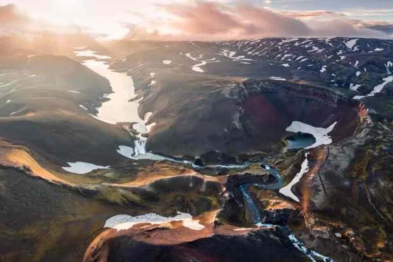 Stunning aerial view of Iceland’s highlands at sunset, featuring winding rivers, snow-capped hills, and rugged volcanic craters under a golden sky.