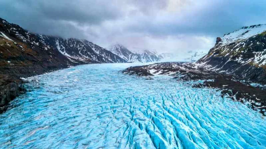 A vast glacier stretches between two mountain ranges, with the icy surface showcasing deep crevices and ridges, under a cloudy sky.