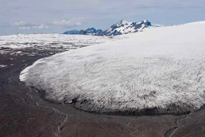 An expansive view of a glacier with a large, ice-covered expanse, surrounded by rugged terrain and distant snow-capped mountains under a clear sky.