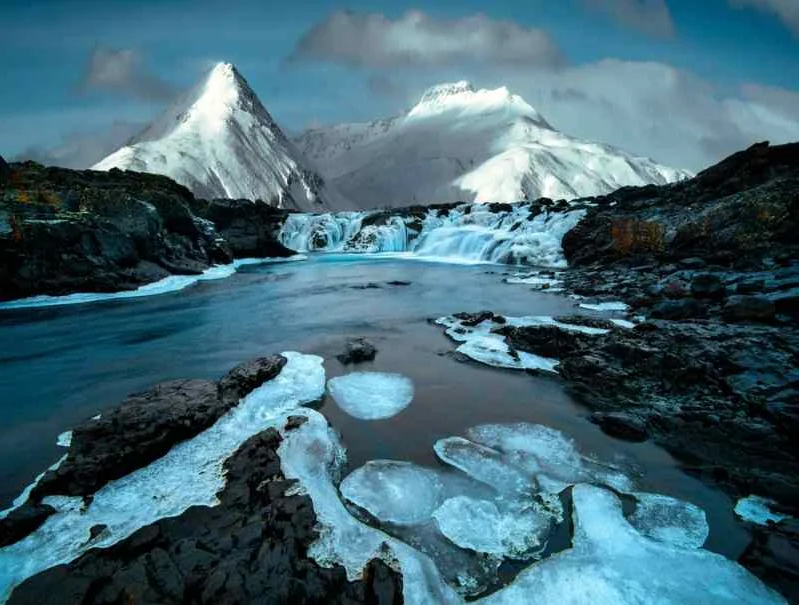 A serene icy river with patches of ice floating on the surface, leading to a waterfall with snow-covered mountains in the background under a cloudy sky.