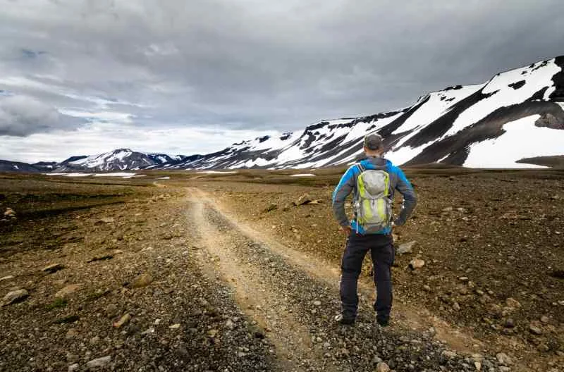 Hiker standing on a rocky trail in a vast, barren landscape with snow-capped mountains in the distance under a cloudy sky.