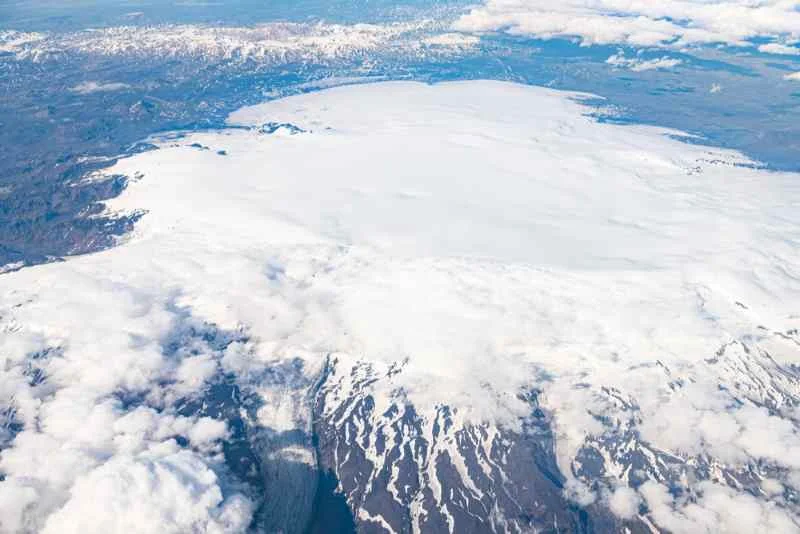 Aerial view of the vast snow-covered Vatnajökull glacier in Iceland, showcasing the rugged mountainous terrain beneath and distant volcanic landscape.