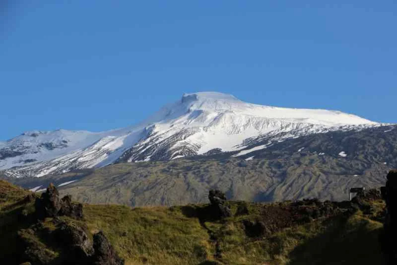 Clear view of Snæfellsjökull volcano in Iceland, with its snow-covered peak rising above green moss-covered lava fields under a bright blue sky.