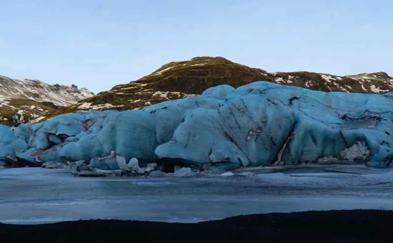 A massive blue glacier with rugged ice formations, set against a backdrop of snow-capped mountains and a clear sky.