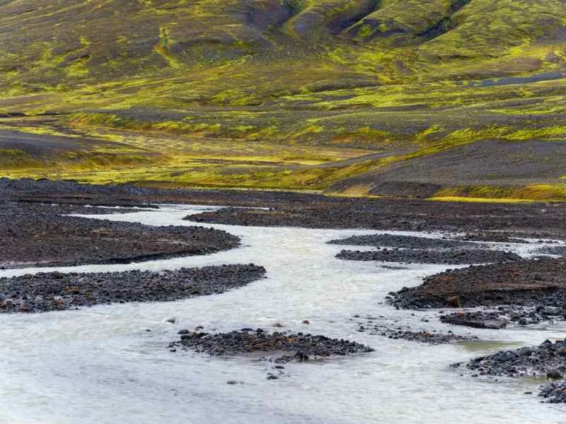 Close-up of a meandering river flowing through a barren, rocky landscape in Iceland, with vibrant green moss covering the surrounding hills.