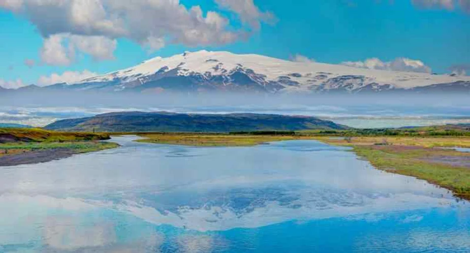 A serene landscape featuring a calm river reflecting the snow-capped mountain in the background under a bright blue sky with scattered clouds.