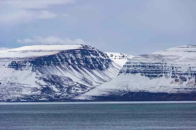 A tranquil seascape with snow-covered mountains rising above the calm water, under an overcast sky.