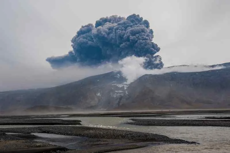 A large volcanic eruption with thick black smoke billowing into the sky from a mountain, with a river flowing in the foreground.