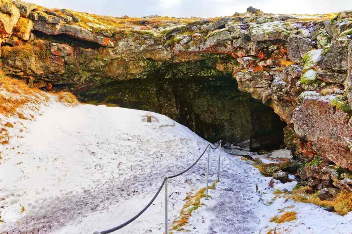 Entrance to a lava cave in Iceland during winter, with snow-covered ground, rugged rock formations, and a path leading into the dark cave.