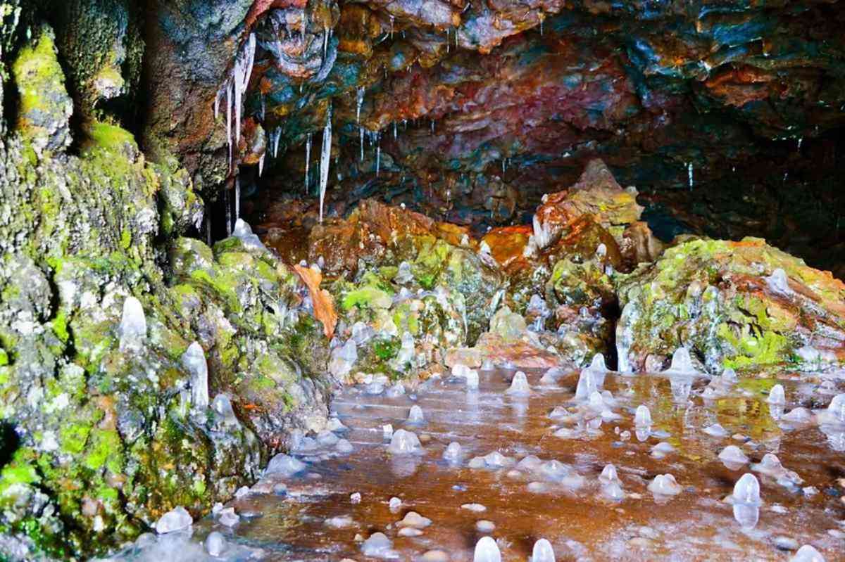 Inside view of a lava cave in Iceland, featuring colorful rock formations, ice stalagmites on the floor, and icicles hanging from the ceiling.