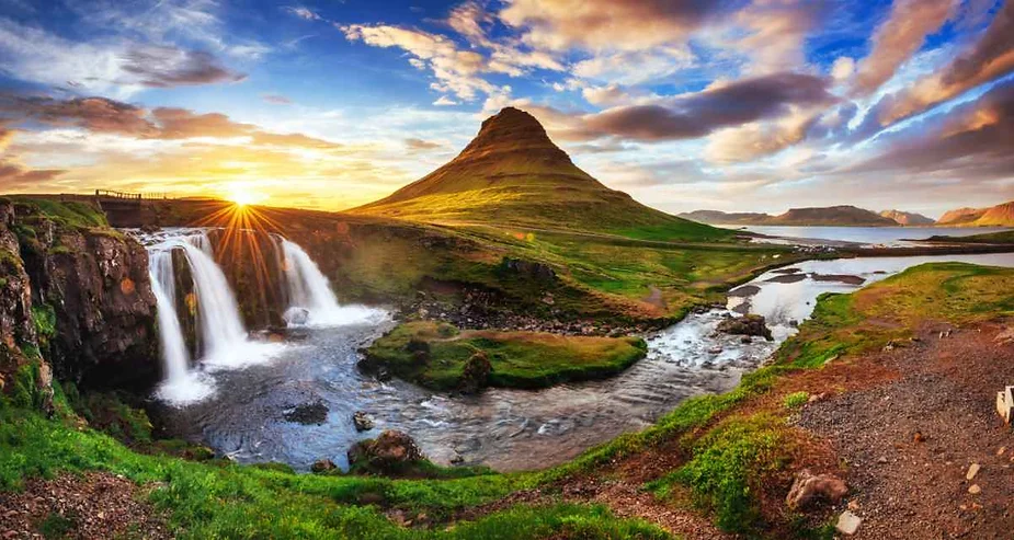 Iconic view of Kirkjufell mountain with the Kirkjufellsfoss waterfall in the foreground during a vibrant sunset in Iceland, with the sun’s rays casting a golden glow on the landscape.