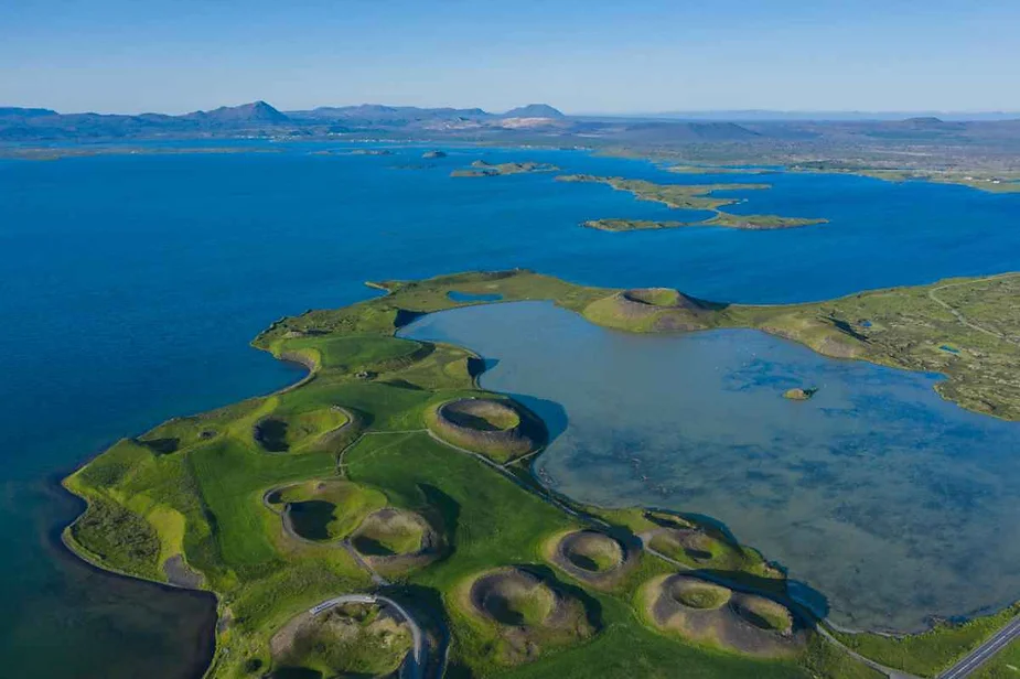 Aerial view of Lake Mývatn in Iceland, featuring unique volcanic craters, lush green landscape, and the expansive blue waters of the lake under a clear sky.