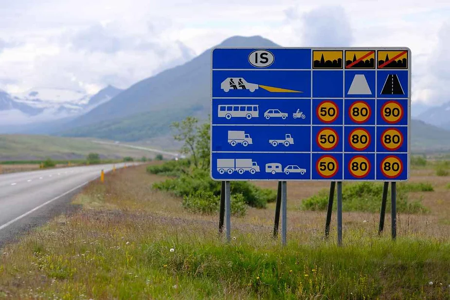 Icelandic road sign displaying speed limits for different types of vehicles in various driving conditions, set against a backdrop of scenic mountains and open road.