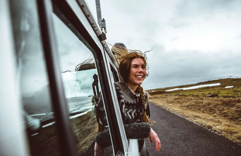 Young woman with a joyful smile leaning out of a moving car’s window on a road trip, with her hair blowing in the wind against a backdrop of Iceland’s rugged landscape.