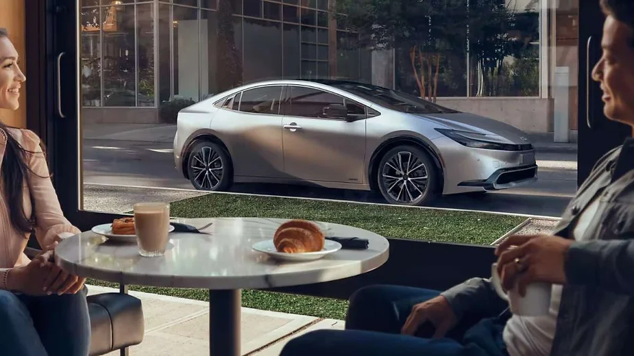 A sleek silver car parked outside a café, viewed through a large window as two people enjoy coffee and croissants inside, blending urban sophistication with everyday life.