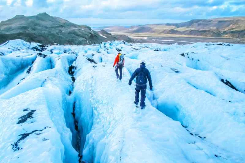 Two hikers equipped with gear walk across a vast glacier in Iceland, navigating deep crevasses and icy terrain, with distant mountains and valleys in the background.