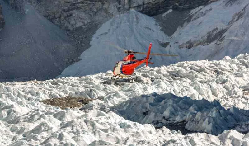 A red helicopter landing on a rugged glacier in Iceland, surrounded by jagged ice formations and steep mountainous terrain in the background.