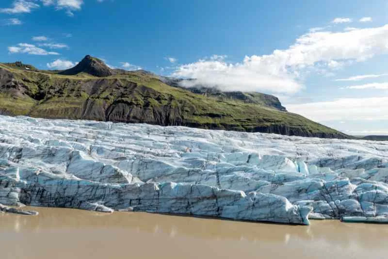 View of a glacier in Iceland with jagged ice formations leading to a muddy glacial lake, set against the backdrop of green mountains under a clear blue sky.