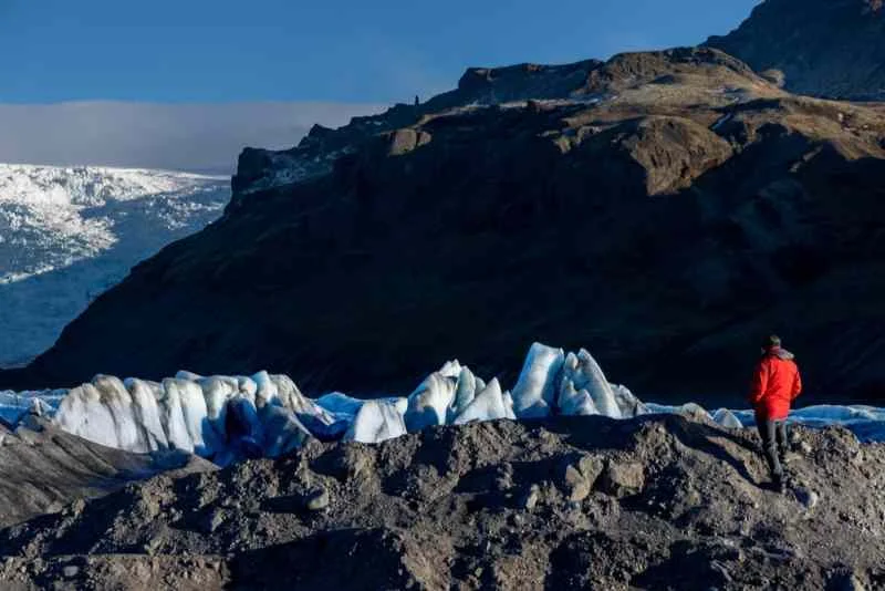 A lone hiker in a red jacket stands on rocky terrain, overlooking jagged blue ice formations of a glacier in Iceland, with towering mountains in the background.