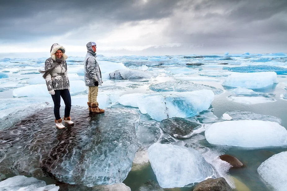 Tourists standing on a large ice floe surrounded by scattered ice chunks at Jökulsárlón Glacier Lagoon in Iceland, dressed in warm winter clothing.