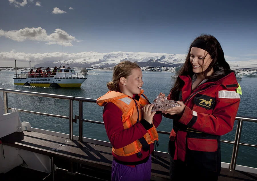 Tourists on a boat tour at Jökulsárlón Glacier Lagoon in Iceland, holding an ice chunk with a backdrop of floating icebergs and snow-covered mountains.