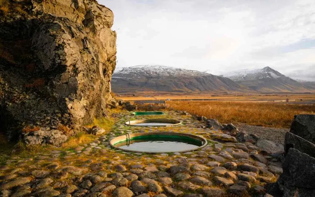 Scenic hot tubs at Hoffell in Iceland, nestled among rocky terrain with a backdrop of snow-capped mountains and golden fields under a soft morning light.