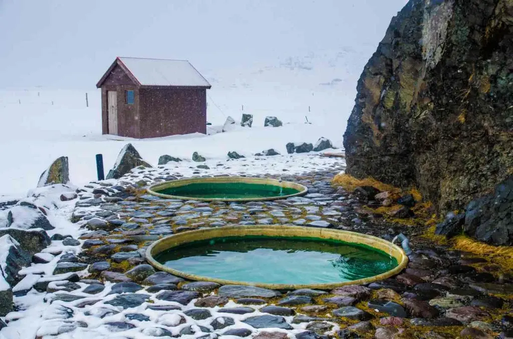 Snow-covered hot tubs at Hoffell in Iceland, surrounded by rocky terrain with a small wooden cabin in the background, creating a cozy winter retreat.