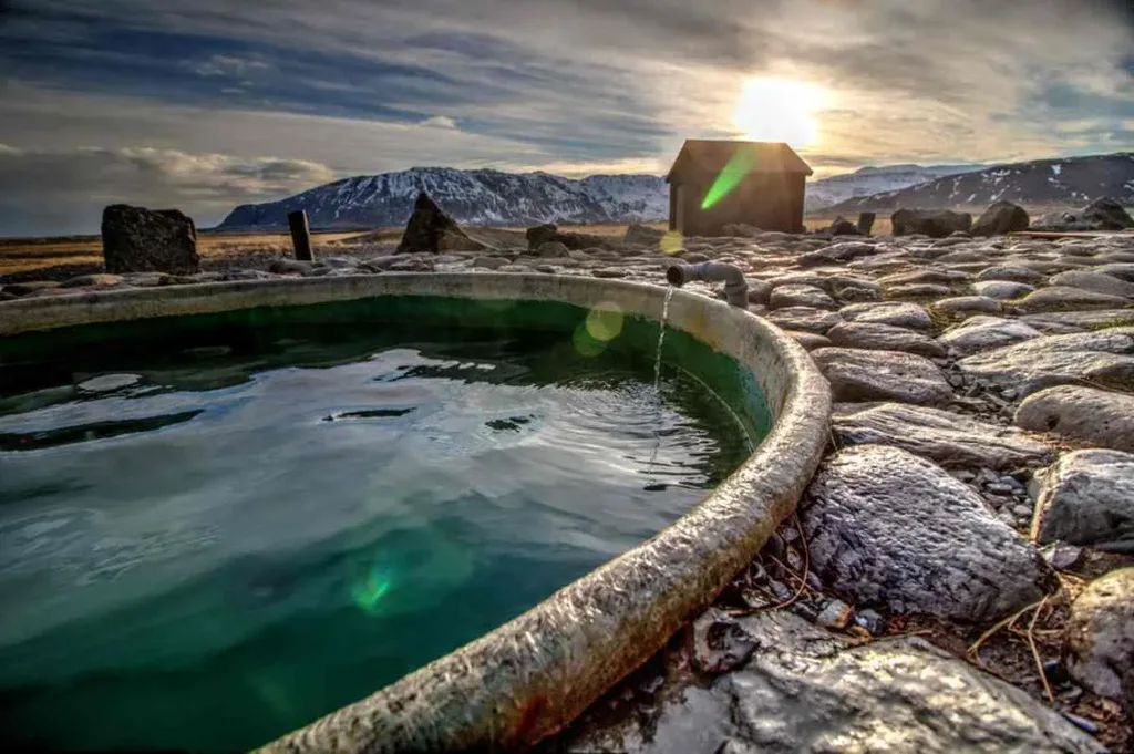 Close-up of a warm Hoffell hot tub in Iceland with rocky surroundings, a small cabin, and a stunning sunset over snow-capped mountains in the distance.