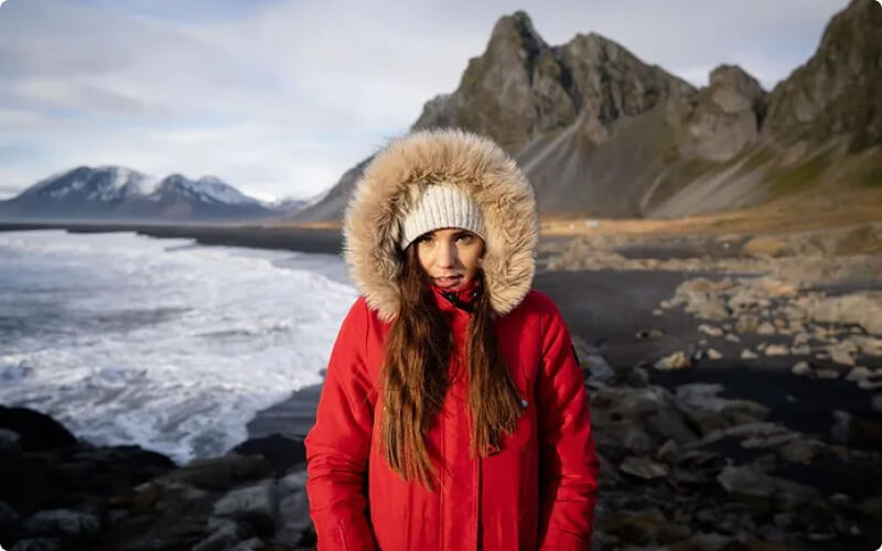 Traveler wearing a red waterproof shell jacket with a fur-lined hood and a knit hat, standing on Iceland’s black sand beach with rugged mountains in the background.