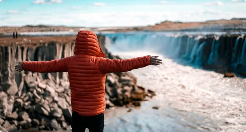 Traveler in a bright red waterproof jacket overlooking a scenic waterfall in Iceland during a rainy fall day, capturing the season's adventure.