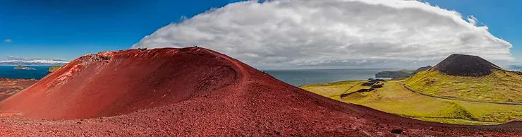 Panoramic view of Eldfell volcano on the Westman Islands in Iceland, featuring vibrant red volcanic slopes and a contrasting green landscape under a cloudy sky.