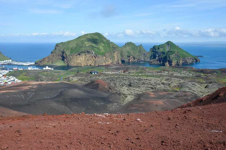 Aerial view of the Westman Islands in Iceland, featuring rugged volcanic terrain, green hills, and the surrounding ocean, seen from a hiking trail.