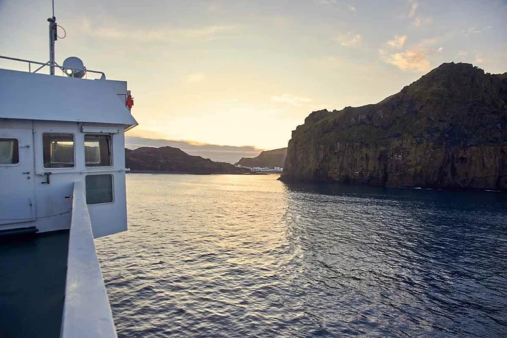 View from the Landeyjahöfn ferry approaching the Westman Islands in Iceland at sunset, with calm waters and rocky cliffs in the background.