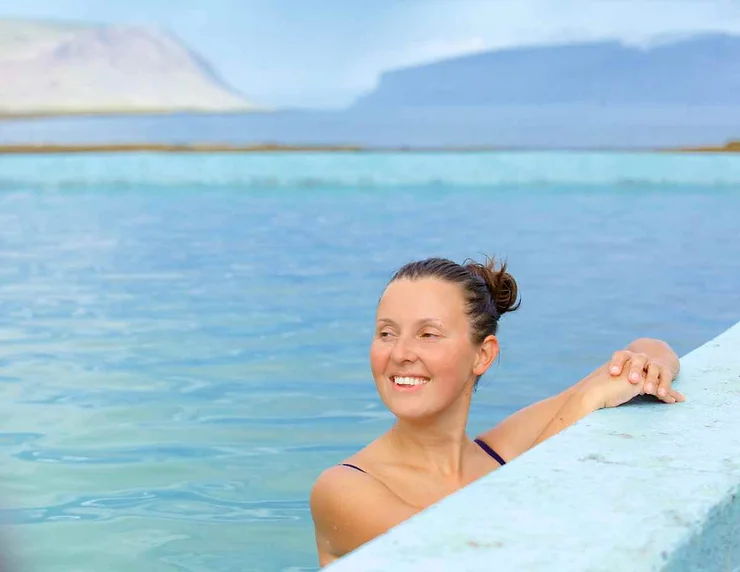 Woman enjoying a relaxing swim in a geothermal pool on the Westman Islands in Iceland, with scenic mountains in the background.