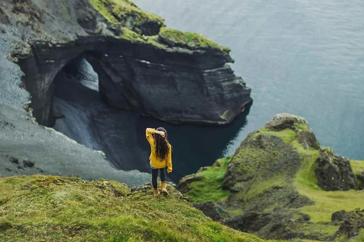 Woman standing on a cliff edge overlooking a dramatic rock formation and ocean on the Westman Islands, Iceland, wearing a yellow jacket.